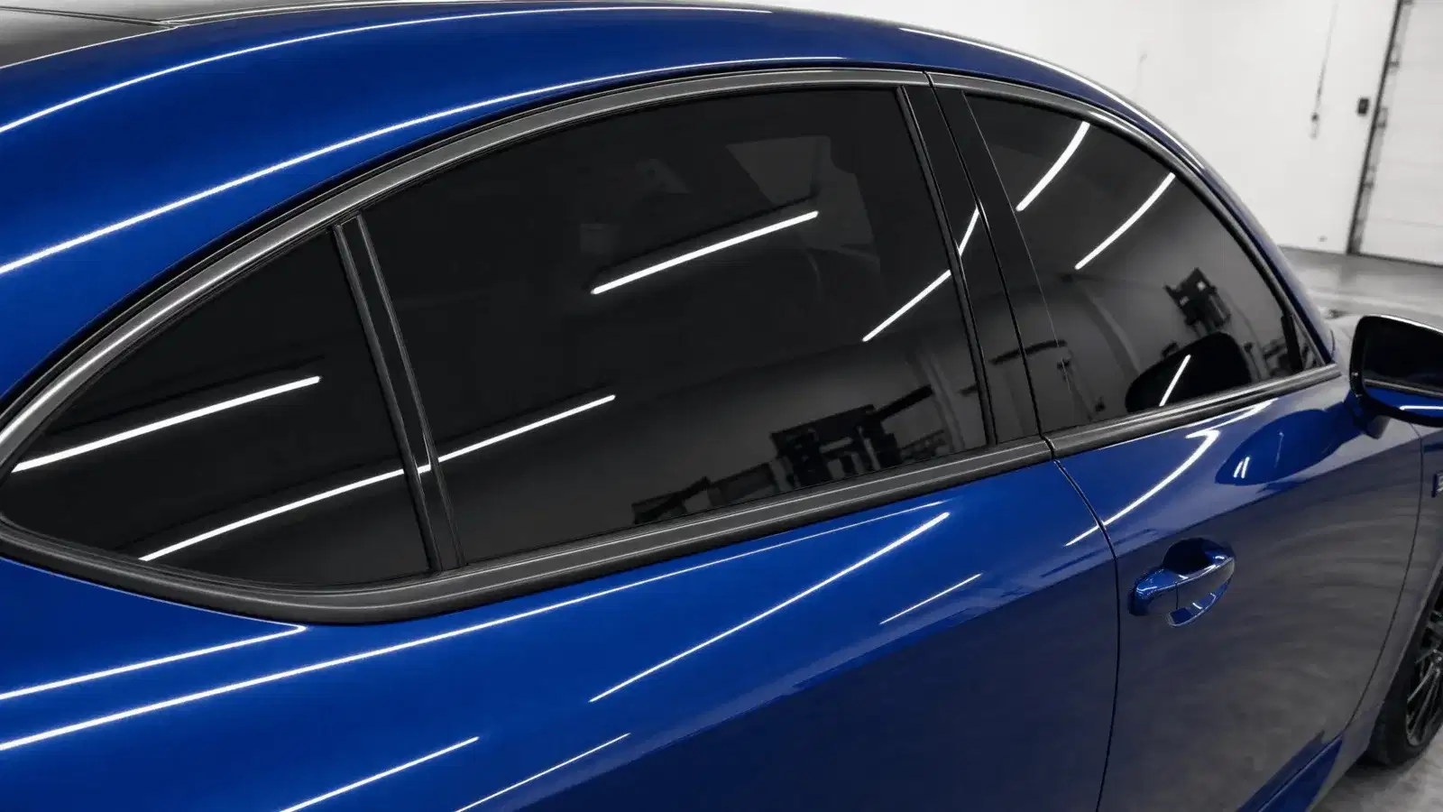 Close-up of black reflective tint on a blue Lexus IS-style sedan inside a clean white tint workshop